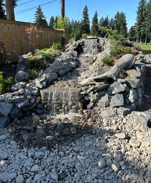 A cascading stone waterfall surrounded by pebbles and greenery, nestled against a wooden fence in a serene outdoor setting.